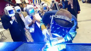 Here's an alt tag for the image: Welder at work with observing audience.